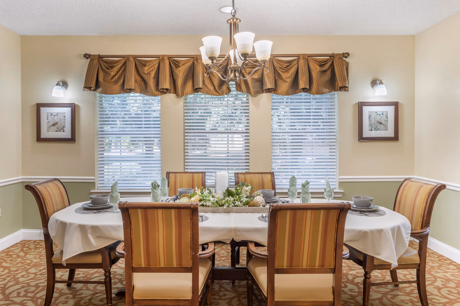 A dining room with a rectangular table covered with a white tablecloth, set with plates, bowls, glasses, and folded green napkins. Six wooden chairs with striped upholstery surround the table. Three windows with white blinds and brown valances are on the wall behind the table. Two framed pictures and wall sconces with lights are on either side of the windows. A chandelier with five lights hangs above the table.