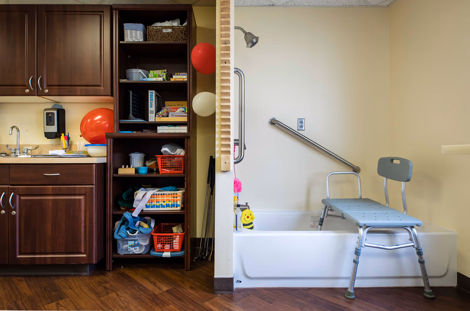 Room interior showing a bathtub with a shower chair and grab bar beside cabinets and open shelving stocked with supplies.