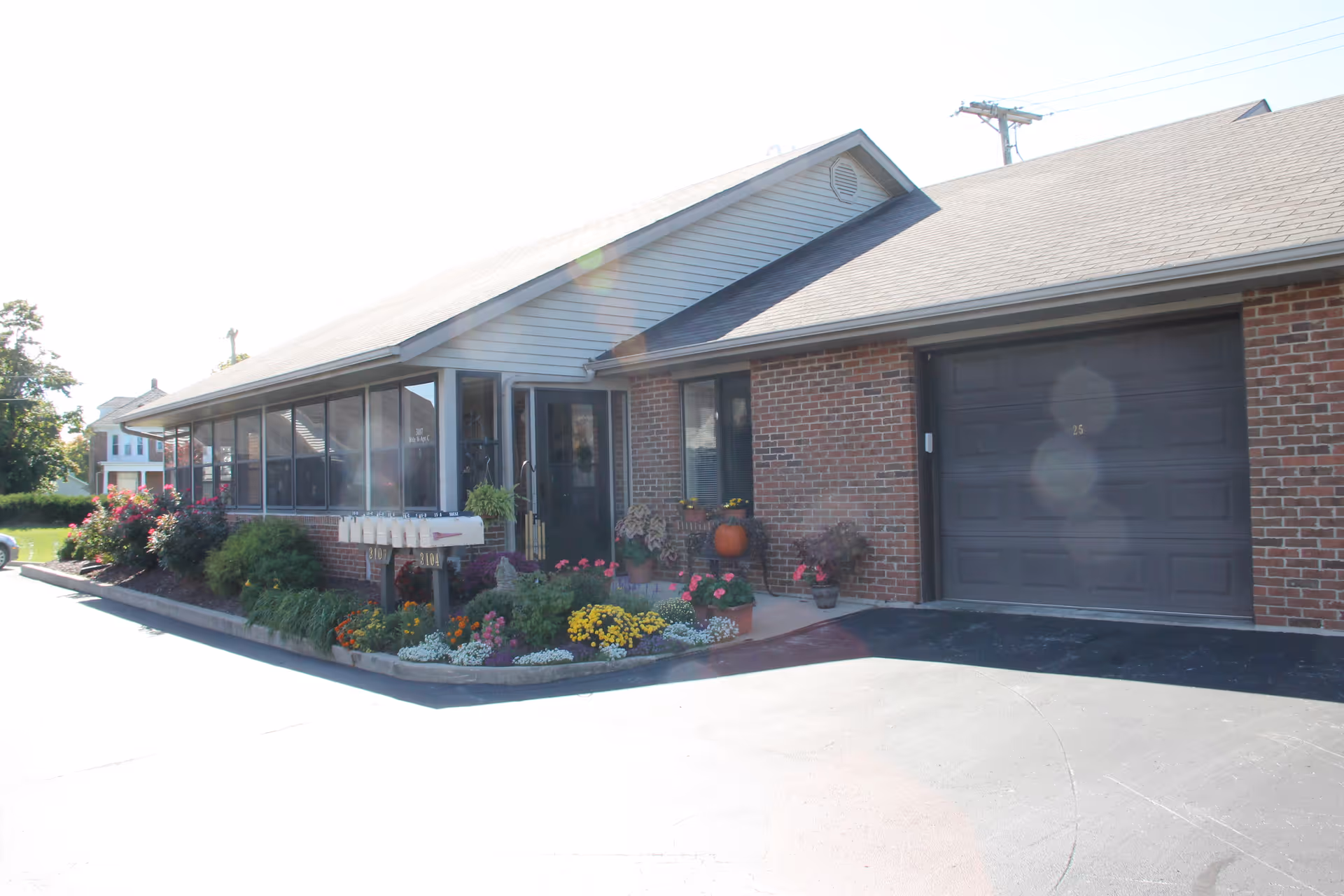 Exterior view of a single-story brick building with a garage door and a screened porch. There are colorful flower beds and shrubs along the front, with a set of mailboxes near the entrance. The building is part of Golden Years Homestead, Inc.