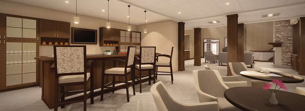 A spacious and well-lit interior common area featuring a bar with high chairs, comfortable armchairs around small tables, and a stone fireplace in the background. The room has wooden pillars, cabinetry, and neutral-toned decor, creating a warm and inviting atmosphere.