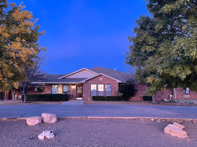 Single-story brick building front with lit windows, trees, and decorative rocks in the foreground under a blue twilight sky.