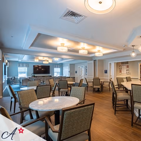 A spacious and well-lit common area with multiple tables and chairs arranged for seating. The room features a large flat-screen TV mounted on the far wall, ceiling lights, and windows letting in natural light. The flooring is wooden, and the walls are painted in neutral tones.