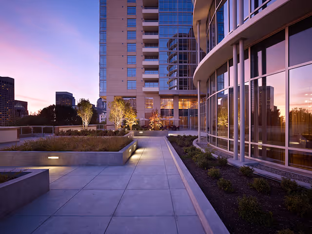 Outdoor patio area at dusk with concrete walkways, raised garden beds with plants, and modern building with large glass windows reflecting the sunset. Trees and city buildings are visible in the background.
