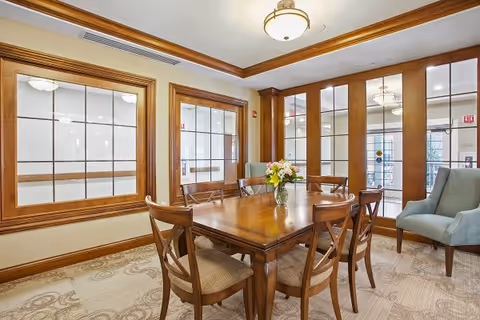 Wood-furnished dining room with a rectangular table, six chairs, a vase of flowers, and paneled interior windows.