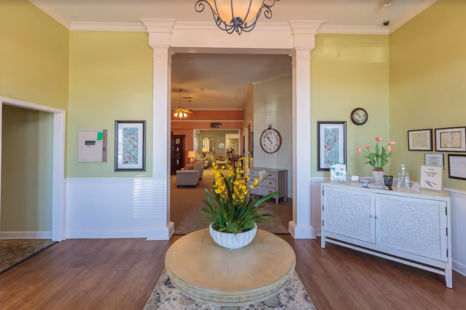 Entry lobby with a round table holding a potted yellow-flowered plant, decorative console and seating area visible through columns.