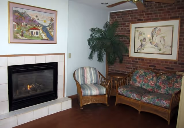 A cozy living room corner with a lit fireplace, wicker chair and loveseat with floral cushions, a potted palm, and framed artwork on a brick accent wall.