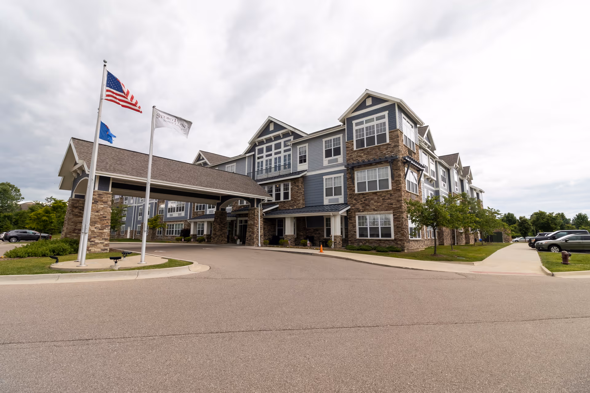 Exterior front view of a multi-story senior living building with a covered entrance, flagpoles, and a driveway.