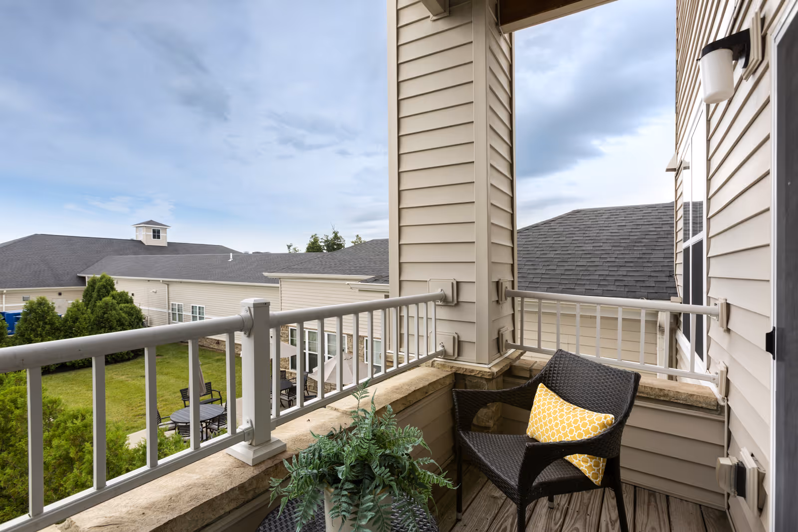 Small furnished balcony with a wicker chair and potted plant overlooking a courtyard and neighboring building roofs.