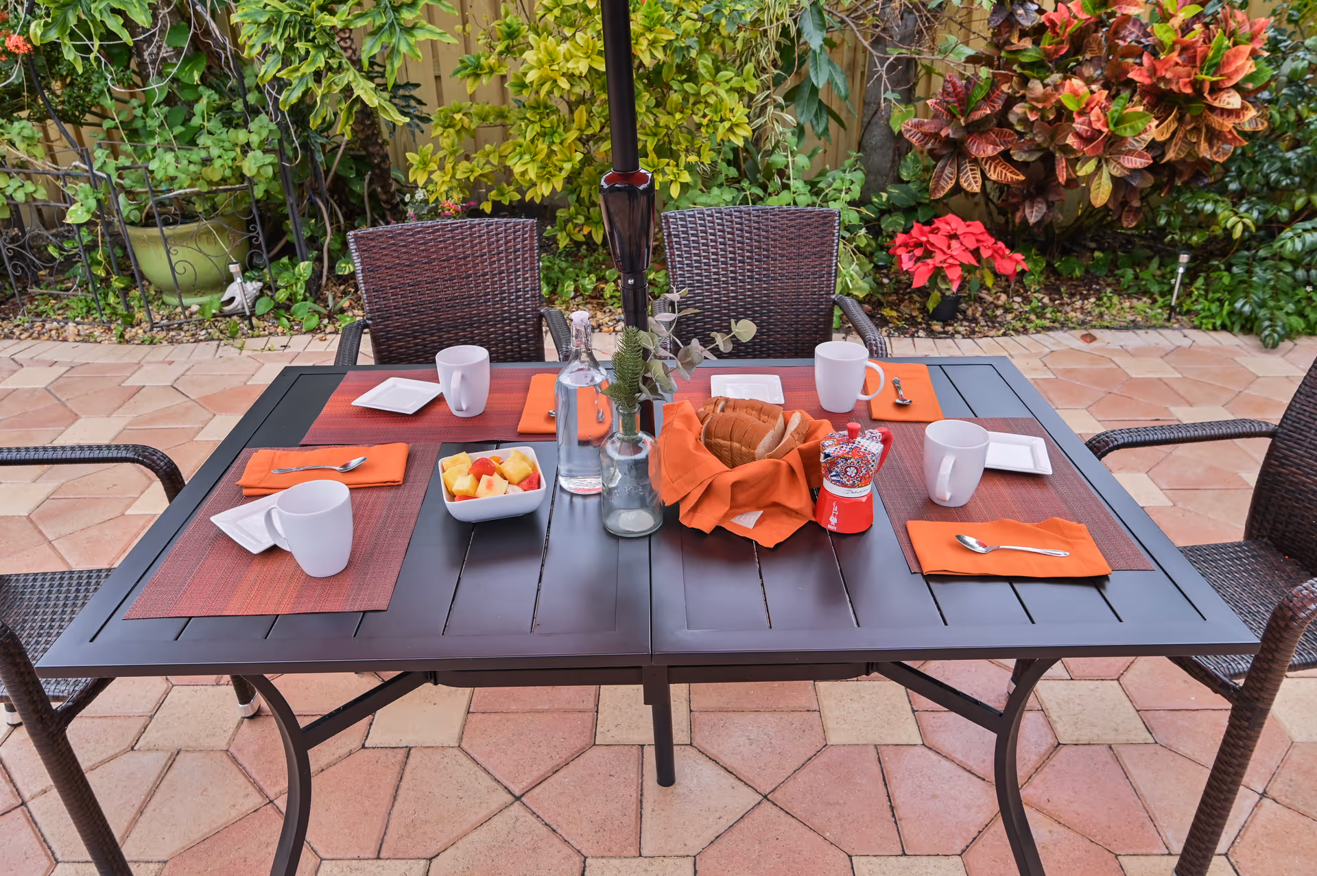 Outdoor patio table set for four with orange placemats, mugs, a fruit bowl and bread basket on a tiled patio with plants in the background.
