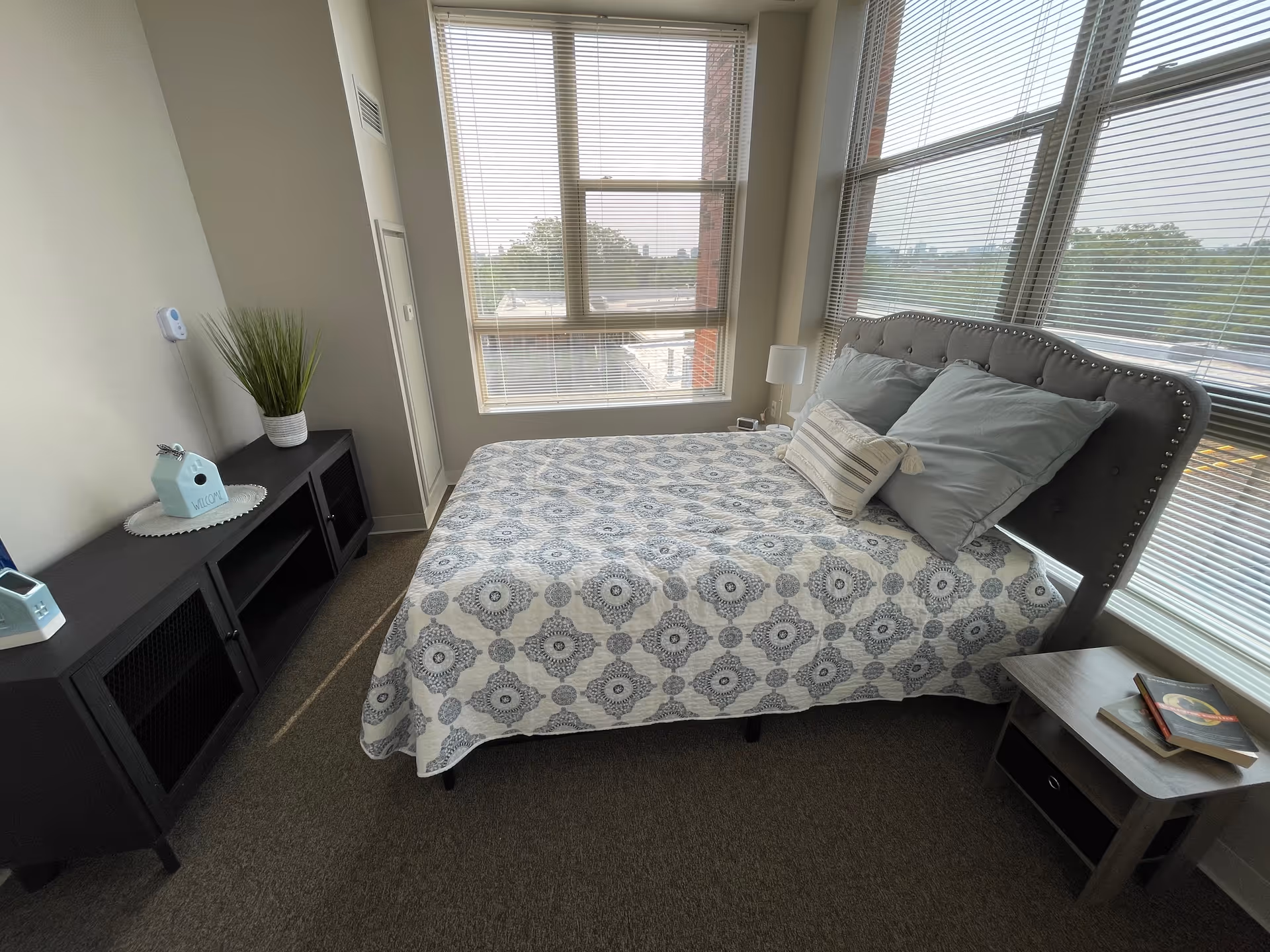 A bright bedroom with large windows covered by blinds. The room features a bed with a gray upholstered headboard, blue and white patterned bedding, and several pillows. There is a small wooden nightstand with books on it next to the bed, and a black cabinet with decorative items including a plant and a small house-shaped ornament on the opposite side. The room has beige walls and carpeted flooring.