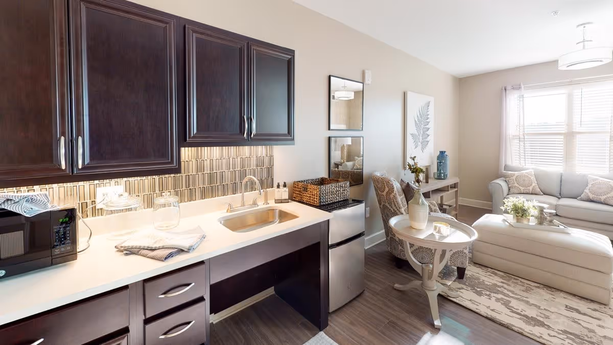 A bright and modern living space featuring a kitchenette with dark wood cabinets, a sink, a microwave, and a mini refrigerator. Adjacent to the kitchenette is a cozy seating area with a patterned armchair, a small round side table, a light-colored sofa, an ottoman, and a window with blinds allowing natural light to fill the room. The walls are decorated with framed artwork and a large botanical print.
