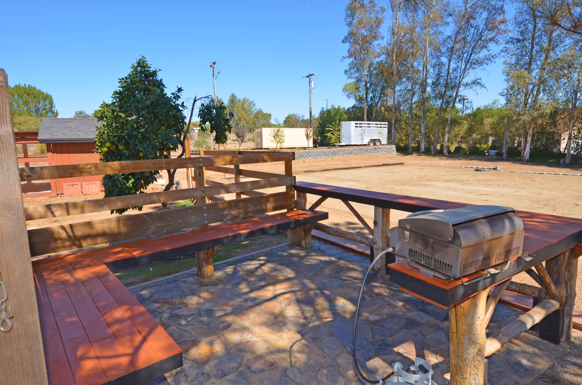 Outdoor seating area with wooden benches arranged in an L-shape around a stone-paved floor. There is a small grill on a wooden stand to the right. In the background, there are trees, a red shed, and an open dirt area under a clear blue sky.