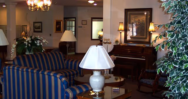 Interior view of a living room area with a blue and gold striped sofa and matching armchair, a white table lamp on a glass coffee table, a piano against the wall with two lamps on either side, framed artwork above the piano, and a large leafy plant on the right side.