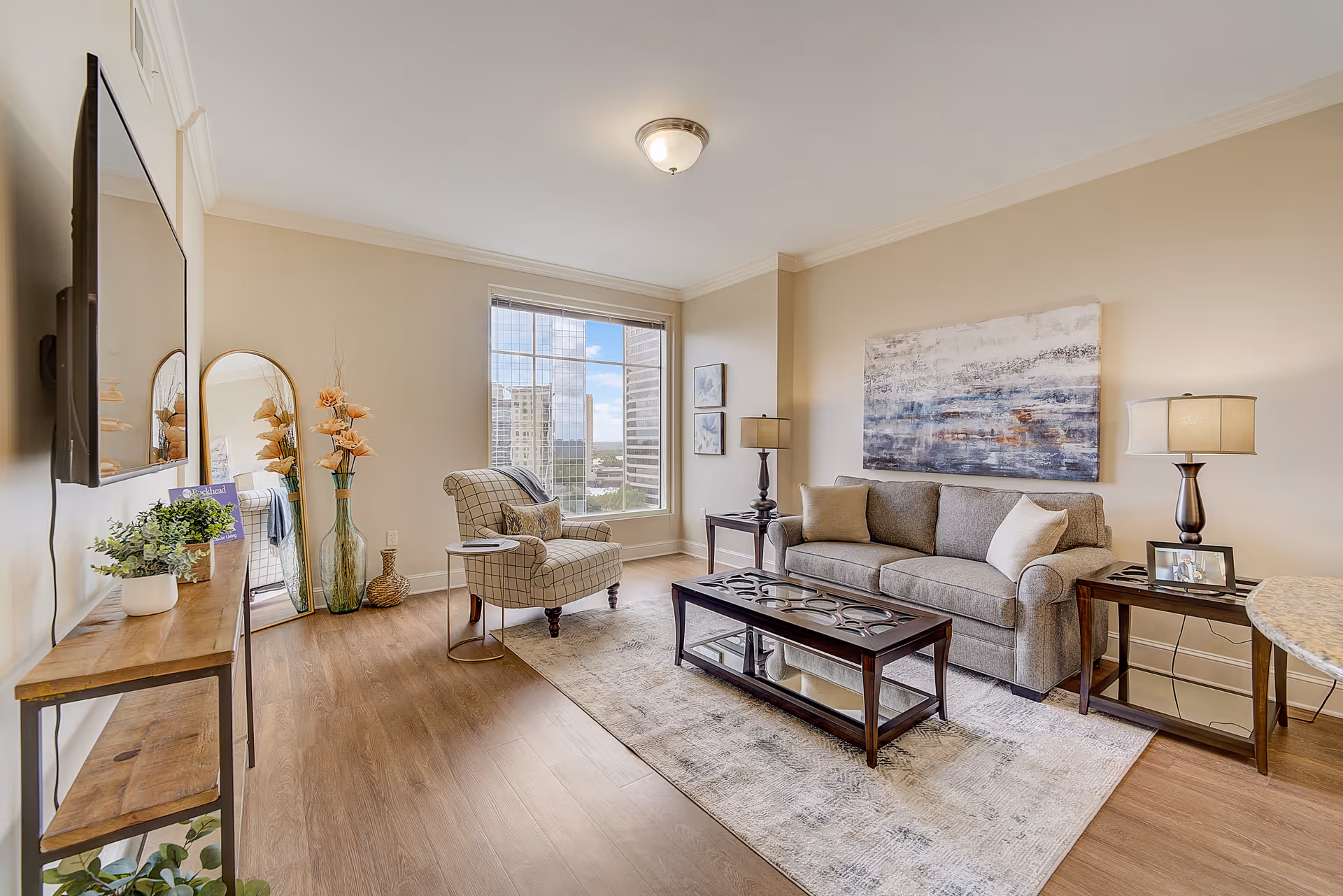 A bright and modern living room with a large window showing a cityscape view. The room features a gray sofa with two pillows, a patterned armchair, a wooden coffee table with a glass shelf, two side tables with lamps, a large abstract painting on the wall, a tall mirror, and decorative vases with flowers. A flat-screen TV is mounted on the wall above a wooden console table with plants and a brochure.