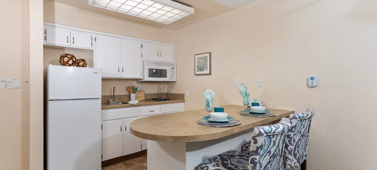 Small kitchenette with white cabinets, refrigerator and microwave, a curved breakfast bar set with place settings and two patterned counter stools.
