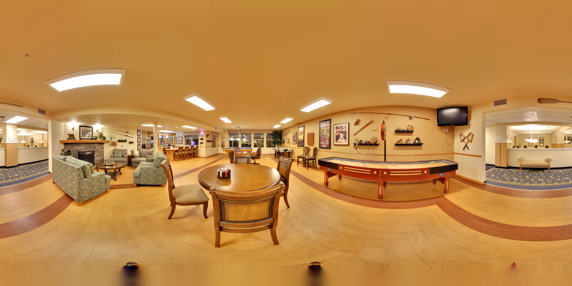 A spacious senior living facility common area featuring a wooden table with chairs in the center, a shuffleboard table against the wall, a seating area with patterned sofas and armchairs near a stone fireplace, and a bar area with stools in the background. The room is warmly lit with ceiling lights and decorated with framed pictures and shelves holding various items. There is a reception desk visible on the right side with a blue carpeted floor area behind it.