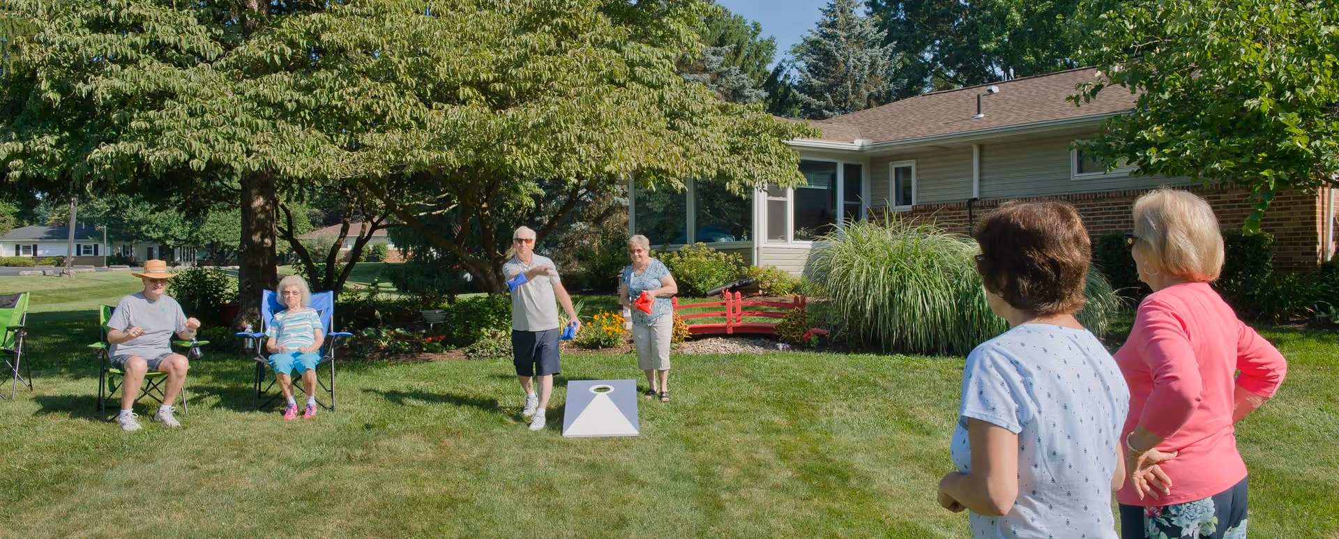 A group of elderly people enjoying a sunny day outdoors at Bethany Village West. Two individuals are seated in lawn chairs under a tree, while three others are standing and playing a game of cornhole on the grass in front of a residential building surrounded by greenery and landscaping.