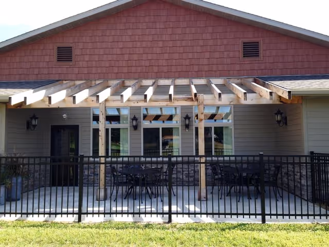 Outdoor patio area of a building with a wooden pergola overhead, black metal fence enclosing the space, and several black metal tables and chairs arranged on a concrete floor. The building exterior features beige siding with stone accents and three wall-mounted lantern-style lights.