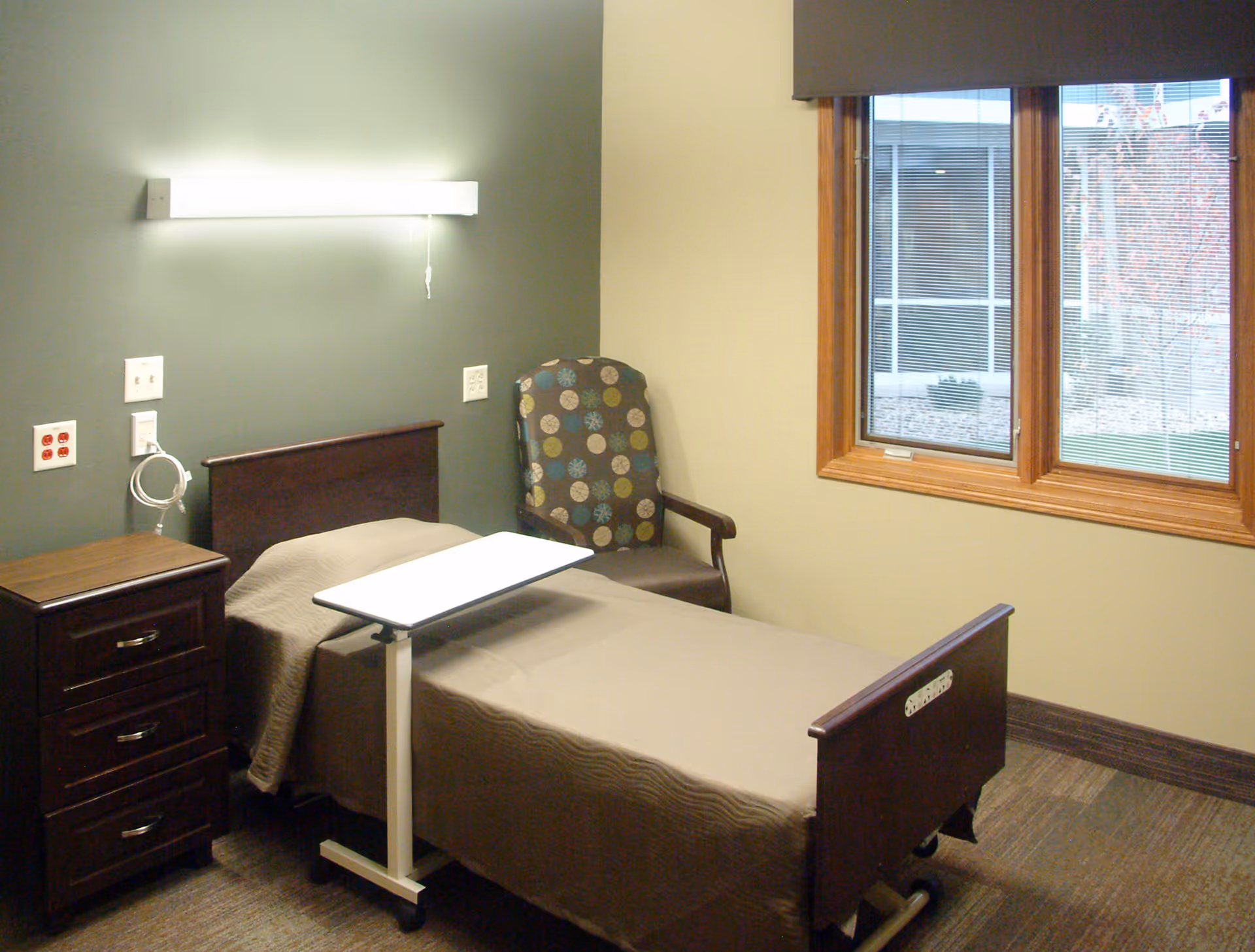 A simple resident bedroom with a single hospital-style bed, overbed table, nightstand, patterned armchair and a window.