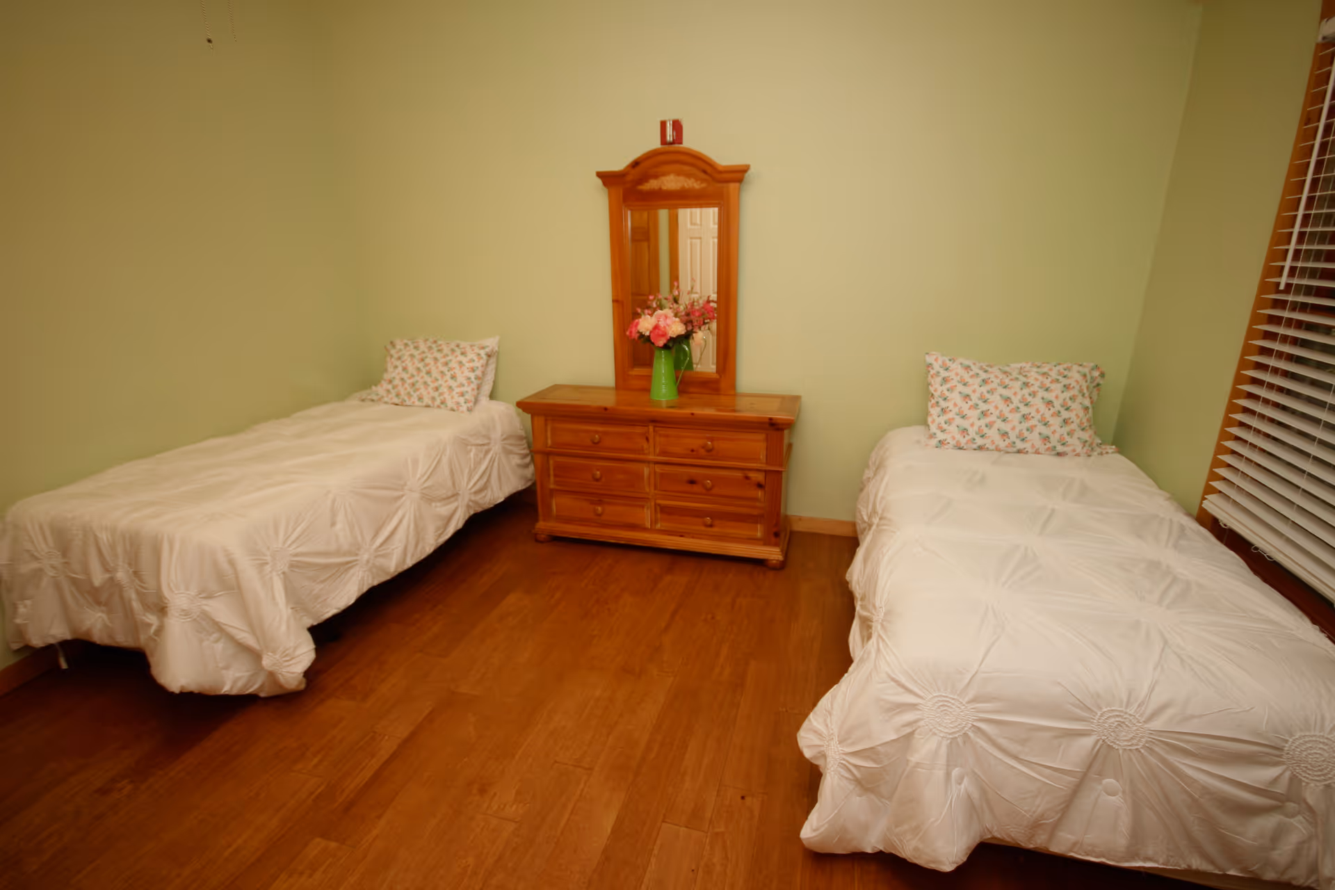A bedroom with two single beds placed against opposite walls, each bed covered with a white textured bedspread and a floral pillow. Between the beds is a wooden dresser with six drawers and a mirror attached. A green vase with pink flowers sits on top of the dresser. The room has light green walls and wooden flooring, with a window featuring wooden blinds on the right side.