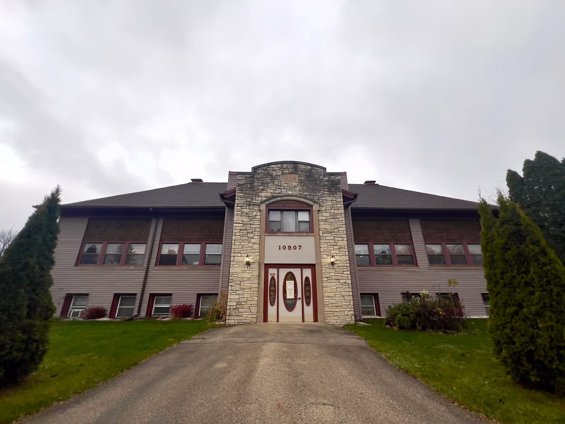 Front exterior view of a two-story building with a stone facade around the entrance. The building has multiple windows with red trim and a dark roof. There is a paved driveway leading up to the entrance, flanked by green grass and tall evergreen shrubs on both sides. The sky is overcast.