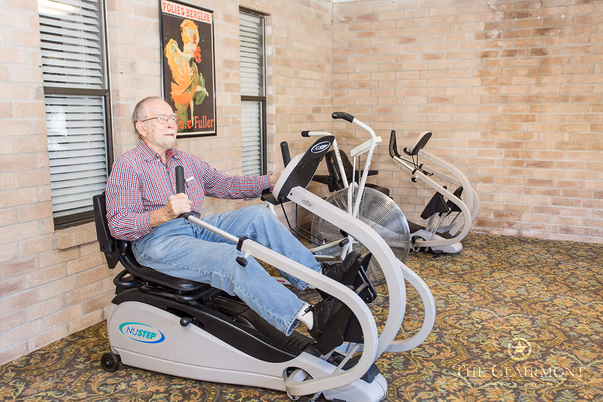 An elderly man wearing a red checkered shirt and blue jeans is exercising on a NuStep recumbent cross trainer in a room with brick walls and patterned carpet. Behind him, there are two other exercise machines and a vintage-style poster on the wall.