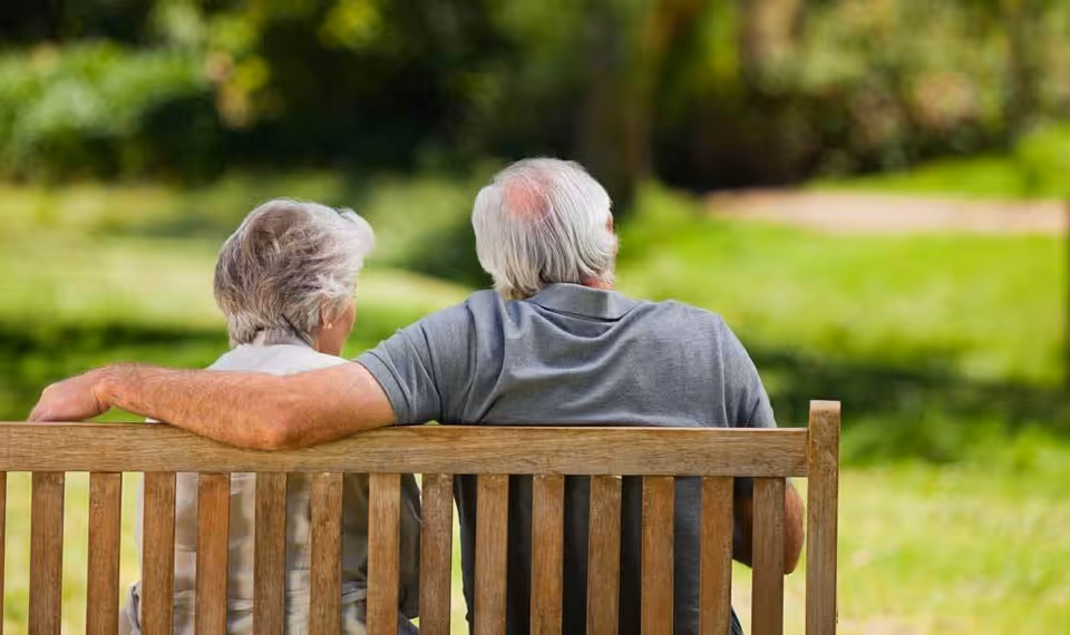 An elderly couple sitting closely together on a wooden bench in a green outdoor park or garden, viewed from behind.