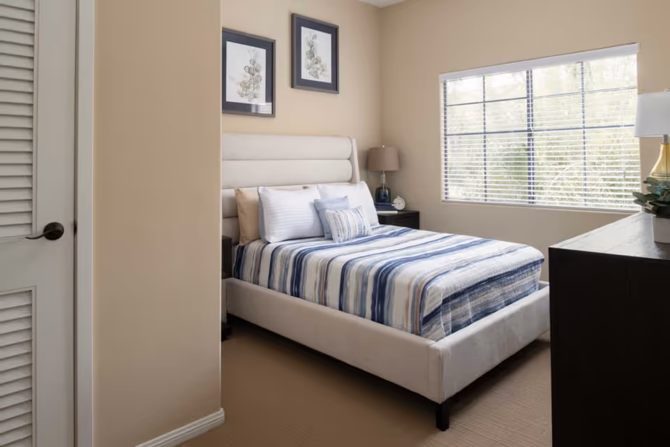 A neatly made bed with a striped blue and white bedspread in a bedroom with beige walls. The bed has multiple pillows and a padded headboard. There are two framed botanical prints hanging above the bed. A window with white blinds lets in natural light. A dark wooden nightstand with a lamp and an alarm clock is beside the bed, and a matching dresser with a lamp and a plant is on the right side of the room.