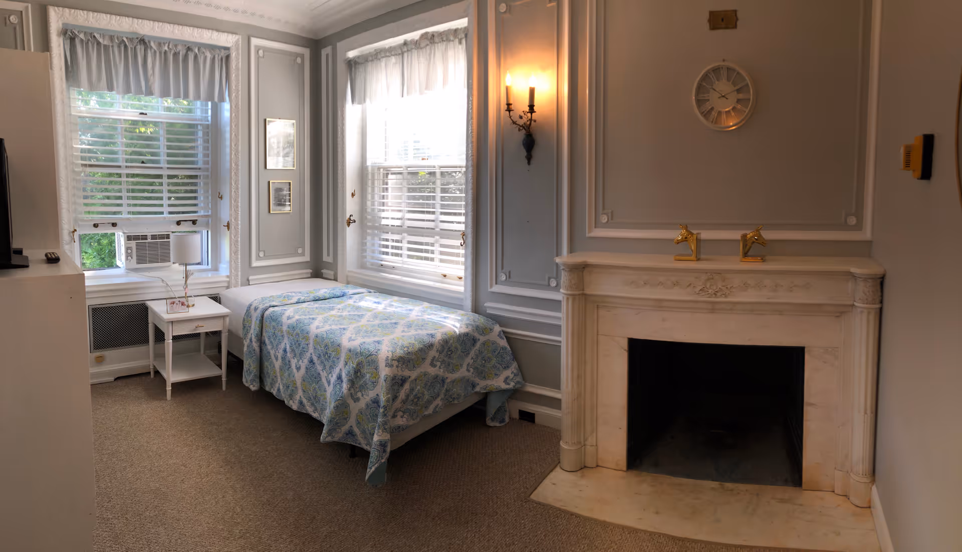 Small bedroom with a single bed by two windows, a white nightstand, and a decorative marble fireplace.