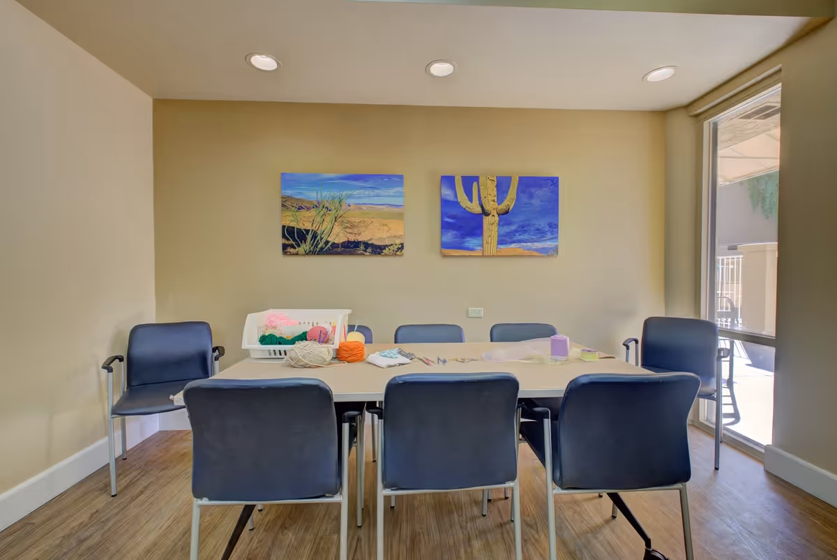 A small activity room with a rectangular table surrounded by six black chairs. On the table, there are various craft supplies including yarn, scissors, and paper. The walls are beige with two colorful desert-themed paintings featuring a cactus and a desert landscape. A large window on the right side lets in natural light and shows an outdoor patio area.