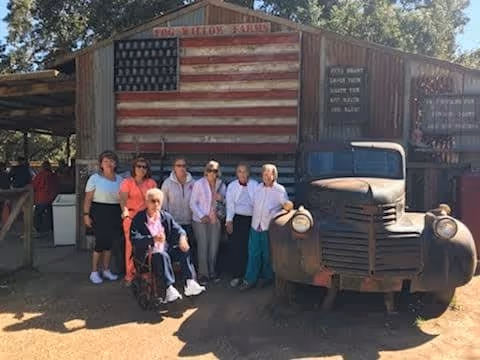 A group of seven elderly people, including one in a wheelchair, posing outdoors in front of a rustic wooden building with a large American flag painted on it. An old rusty truck is parked to the right of the group. Trees and sunlight are visible in the background.