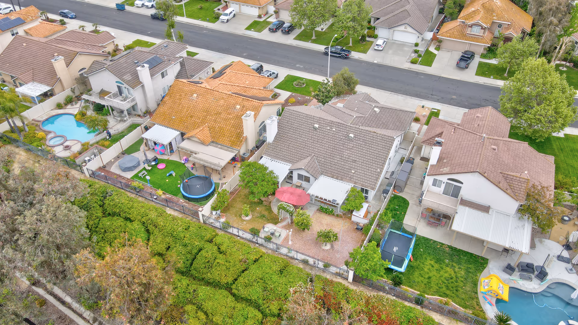 Aerial view of a residential neighborhood showing several houses with backyards. The backyards feature various amenities including a swimming pool, trampoline, patio furniture, and green lawns. The houses have tiled roofs and are separated by fences. A street with parked cars runs parallel to the houses.