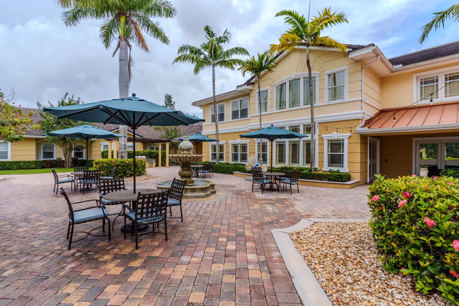 Outdoor courtyard area at Addington Place of Stuart featuring brick-paved ground, several tables with chairs and blue umbrellas, a central water fountain, palm trees, and a yellow two-story building with white-trimmed windows in the background.