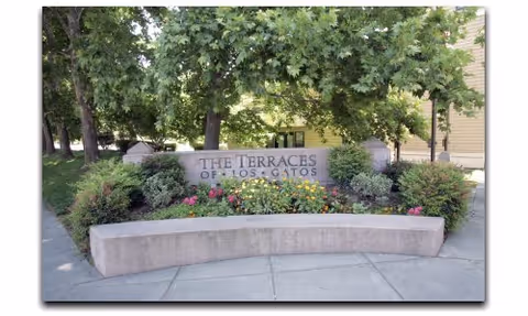 Stone sign reading 'The Terraces of Los Gatos' surrounded by green bushes and colorful flowers, with trees and a building partially visible in the background.
