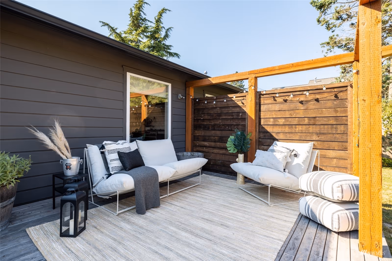 Cozy outdoor patio with white cushioned seating, a wooden privacy fence with string lights, and a striped rug on a wooden deck.