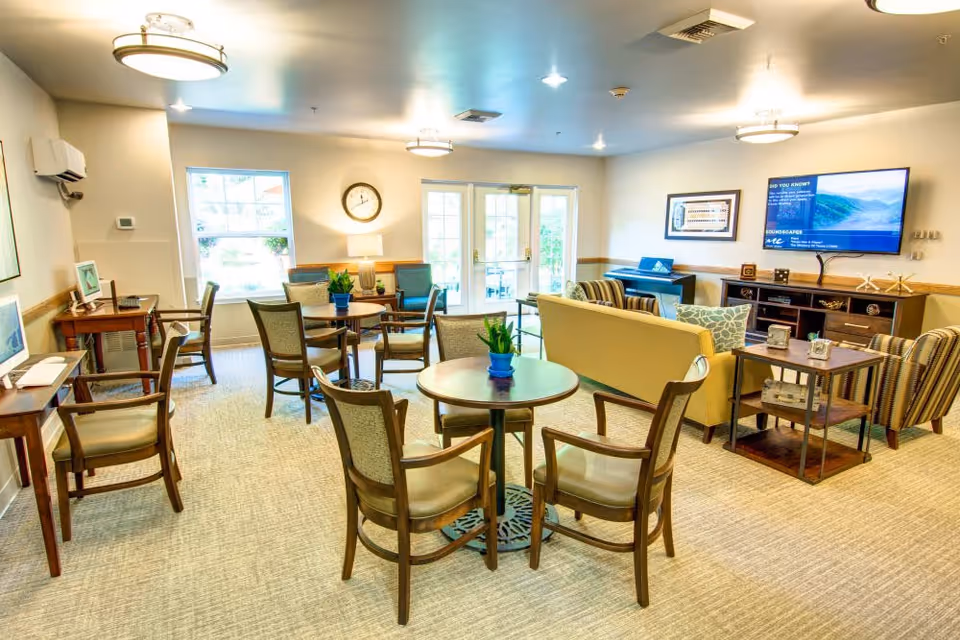 A bright and spacious common area in Bridgeport Place Assisted Living featuring multiple round tables with chairs, a beige sofa, striped armchairs, a wall-mounted TV, a clock, and large windows letting in natural light.