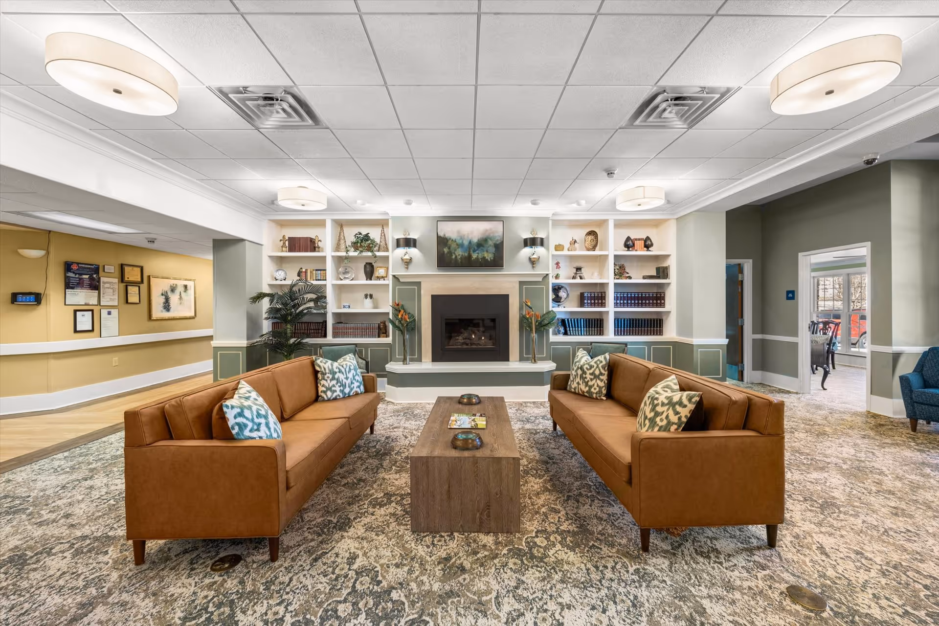A cozy living room area in Windsor Ridge featuring two brown leather sofas facing each other with patterned throw pillows. Between them is a wooden coffee table with decorative items. The room has a patterned carpet and a fireplace centered on the far wall, flanked by built-in white shelves filled with books, plants, and decorative objects. The walls are painted in soft green and yellow tones, and the ceiling has recessed lighting fixtures.