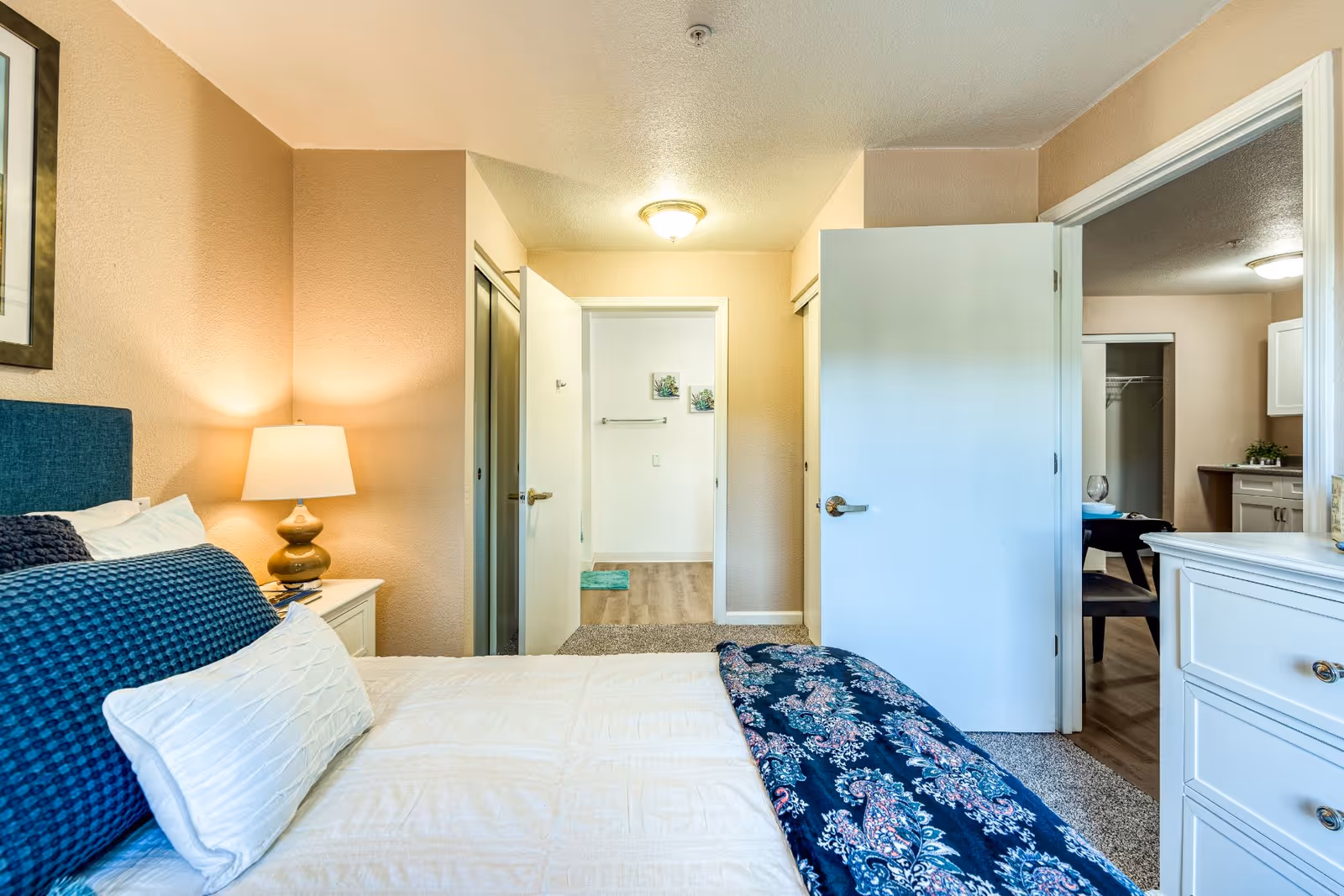 A bedroom in a senior living facility with a neatly made bed featuring white and blue bedding. A bedside table with a lamp is on the left side. The room has beige walls and carpeted flooring. Two open doors reveal a hallway and an adjacent room with a dining table and kitchen area visible.