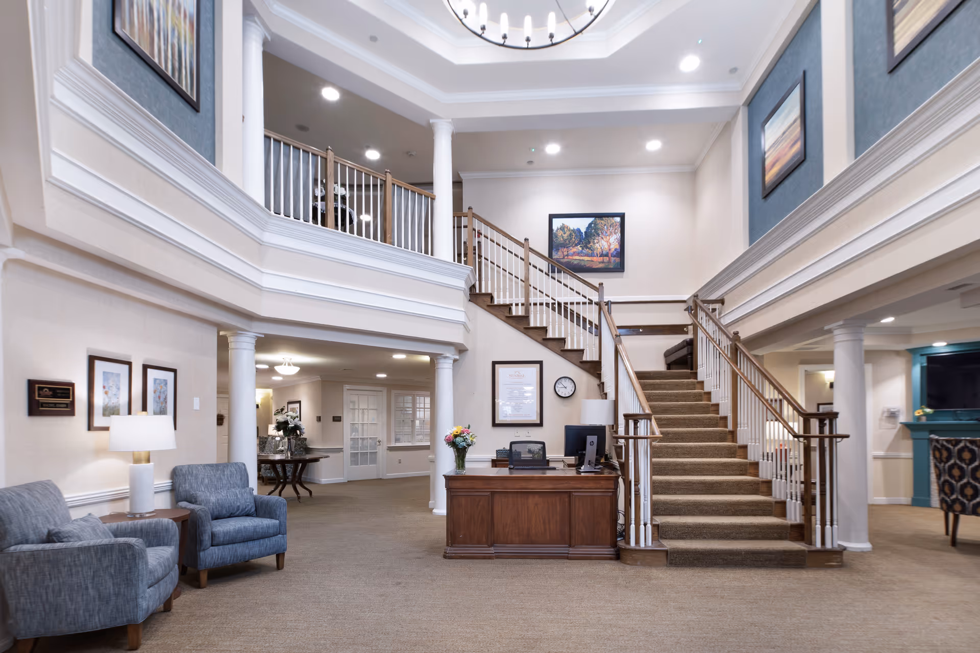 Interior view of a senior living facility lobby with a central wooden reception desk, carpeted staircase leading to an upper floor, blue upholstered chairs, framed artwork on the walls, and white columns supporting the upper balcony.