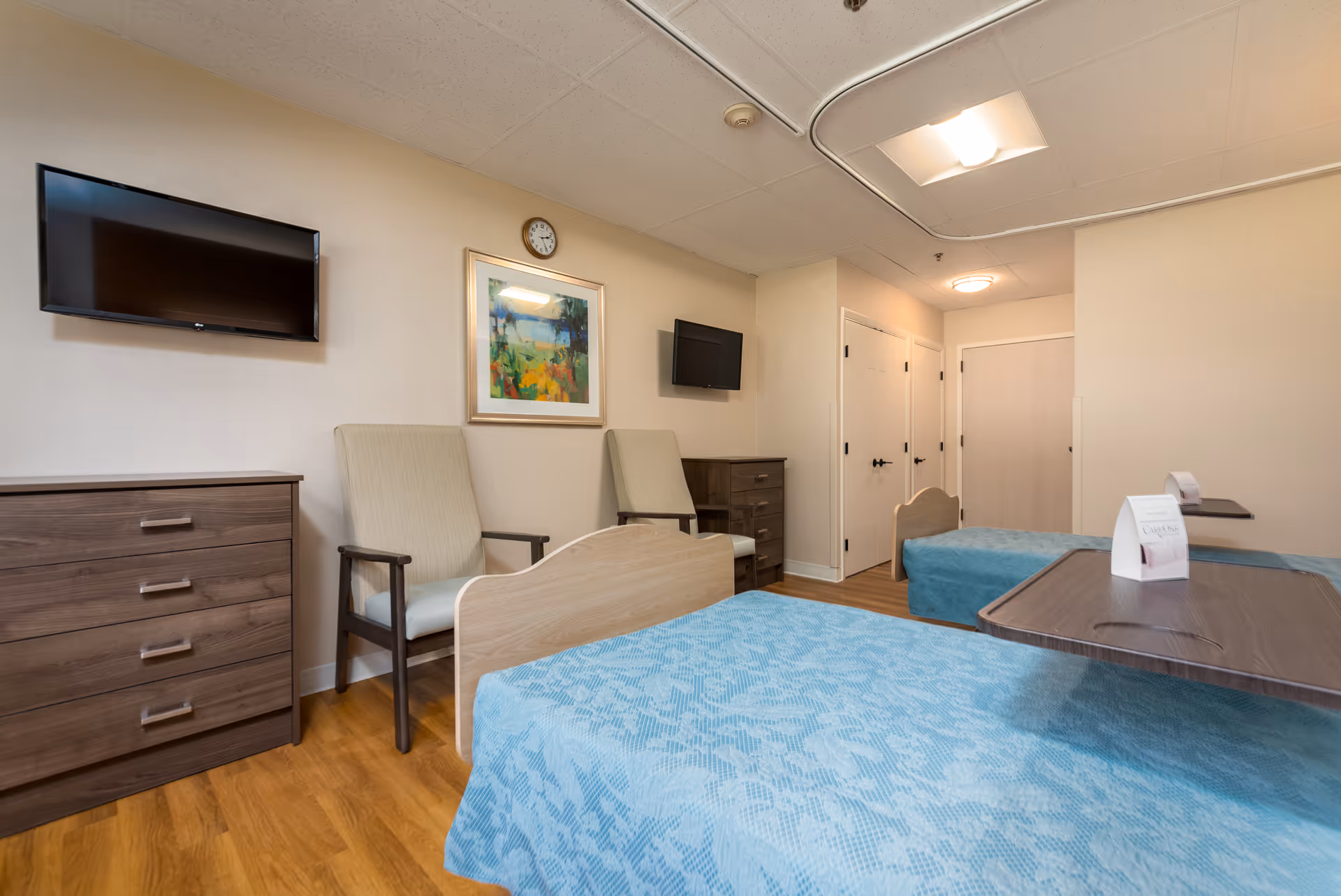 A senior living facility bedroom with two beds covered in blue patterned bedspreads. The room has two beige armchairs, two wall-mounted flat-screen TVs, a wooden dresser, and a framed colorful painting on the wall. The floor is wood, and there are multiple doors visible in the background.