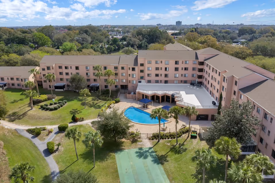 Aerial view of a large senior living facility named Spring Haven with a central swimming pool, surrounded by a multi-story building and landscaped gardens with palm trees and walking paths under a partly cloudy sky.