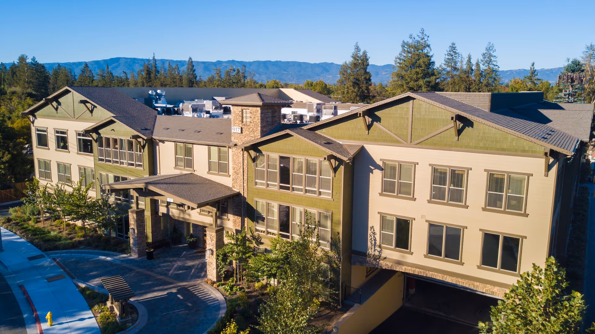 Exterior view of Oakmont of San Jose, a multi-story senior living facility with beige and green walls, multiple windows, a covered entrance, and surrounding trees and landscaping under a clear blue sky with mountains in the background.