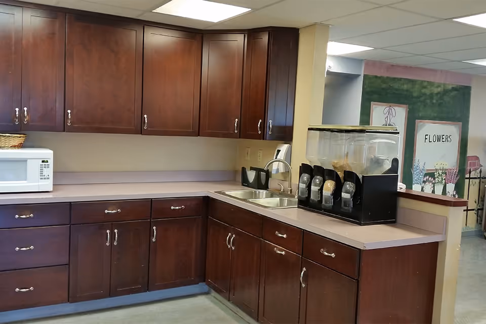 Interior view of a kitchenette area with dark wood cabinets, a microwave on the counter, a double sink, and a cereal dispenser. In the background, there is a wall with a mural featuring flowers and the word 'FLOWERS'.