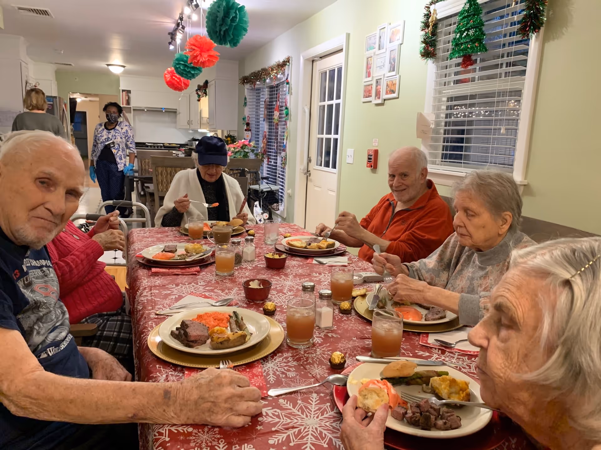 A group of elderly people sitting around a dining table enjoying a meal together in a decorated room with Christmas ornaments and garlands. There are plates of food and glasses of juice on the table, and two staff members are visible in the background near the kitchen area.