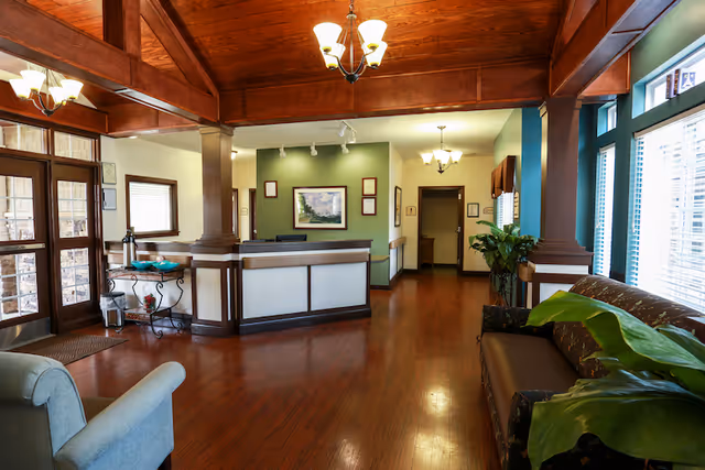 Interior view of a senior living facility lobby with wooden floors and ceiling beams. There is a reception desk in the center with framed pictures on the wall behind it. To the right, there is a brown couch with green plants nearby and large windows letting in natural light. To the left, there is a light blue armchair and a small table with decorative items near the entrance doors.