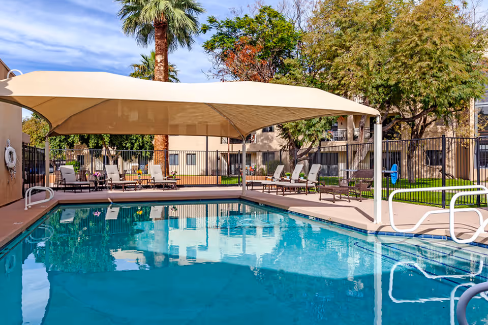 Outdoor swimming pool area with a large beige shade canopy covering several lounge chairs and tables. The pool is surrounded by a fence, and there are trees and a building in the background under a partly cloudy sky.