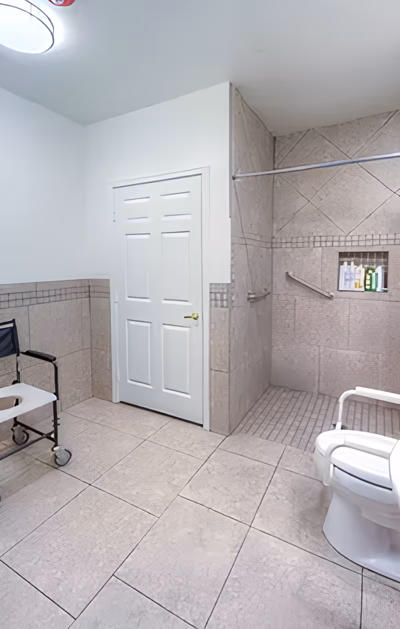 A spacious bathroom with beige tiled floors and walls. There is a white door with a gold handle, a white toilet with armrests, a black shower chair with wheels, and a walk-in shower area with a built-in niche holding toiletries. The shower has a metal grab bar and a curtain rod but no curtain.
