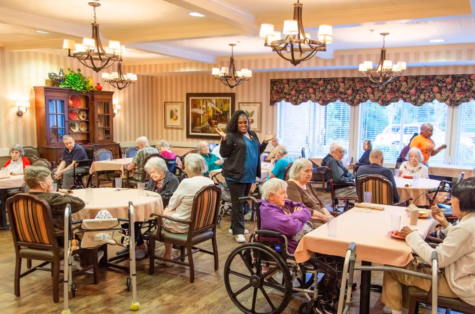 A dining room in Colonial Gardens Assisted Living & Memory Care with elderly residents seated at tables covered with peach-colored tablecloths. A staff member stands smiling in the center of the room. The room is warmly lit with chandeliers and wall sconces, decorated with framed artwork and a floral valance over large windows.