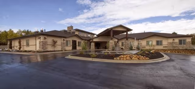 Front exterior of a low-rise senior living building with a covered entry, circular driveway, and landscaped beds under a cloudy sky.