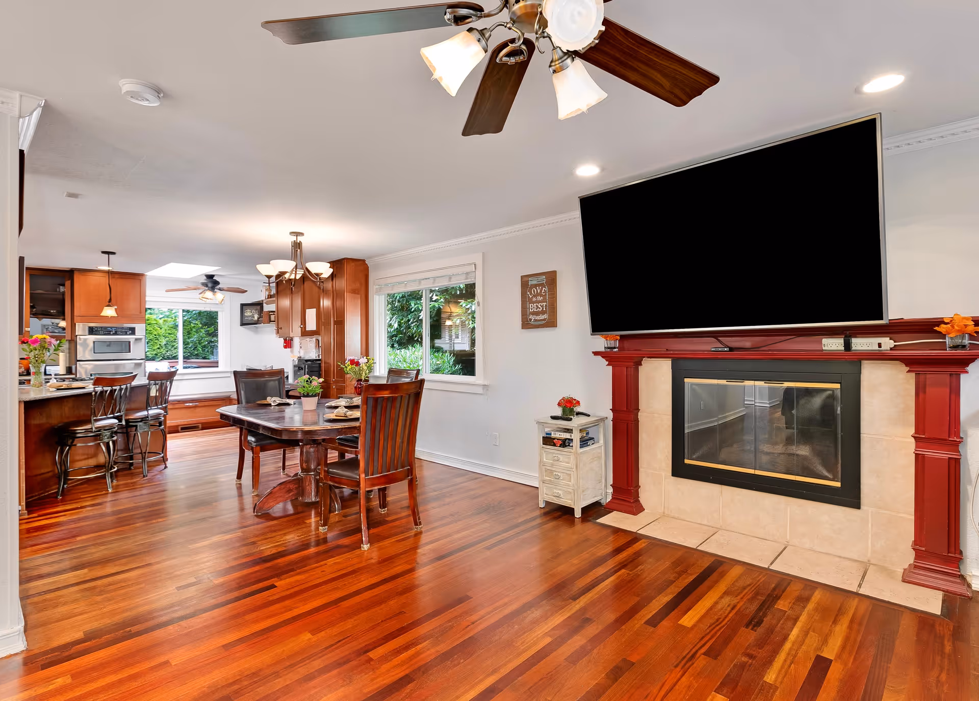 A bright and spacious interior of a senior care home featuring a wooden dining table with chairs, a kitchen area with bar stools, a ceiling fan with lights, a large flat-screen TV mounted above a fireplace with red trim, and a window showing greenery outside.